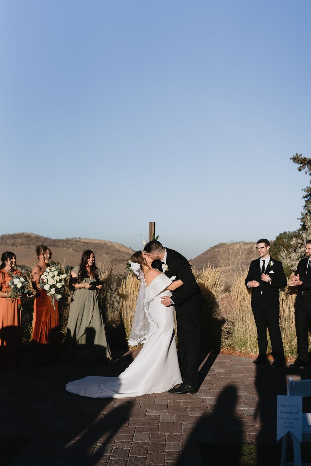 Bride and groom share first kiss at outdoor wedding ceremony with bridesmaids in earth tones and scenic mountain backdrop.