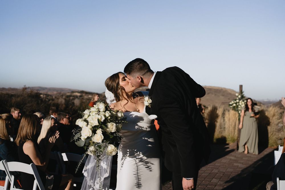 A couple shares their first kiss as newlyweds during an outdoor wedding ceremony with guests seated nearby.