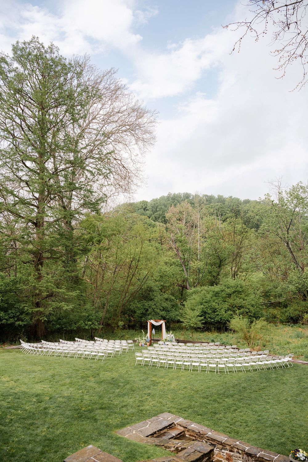 Outdoor wedding ceremony setup with white chairs on green lawn surrounded by lush forest trees under cloudy sky.