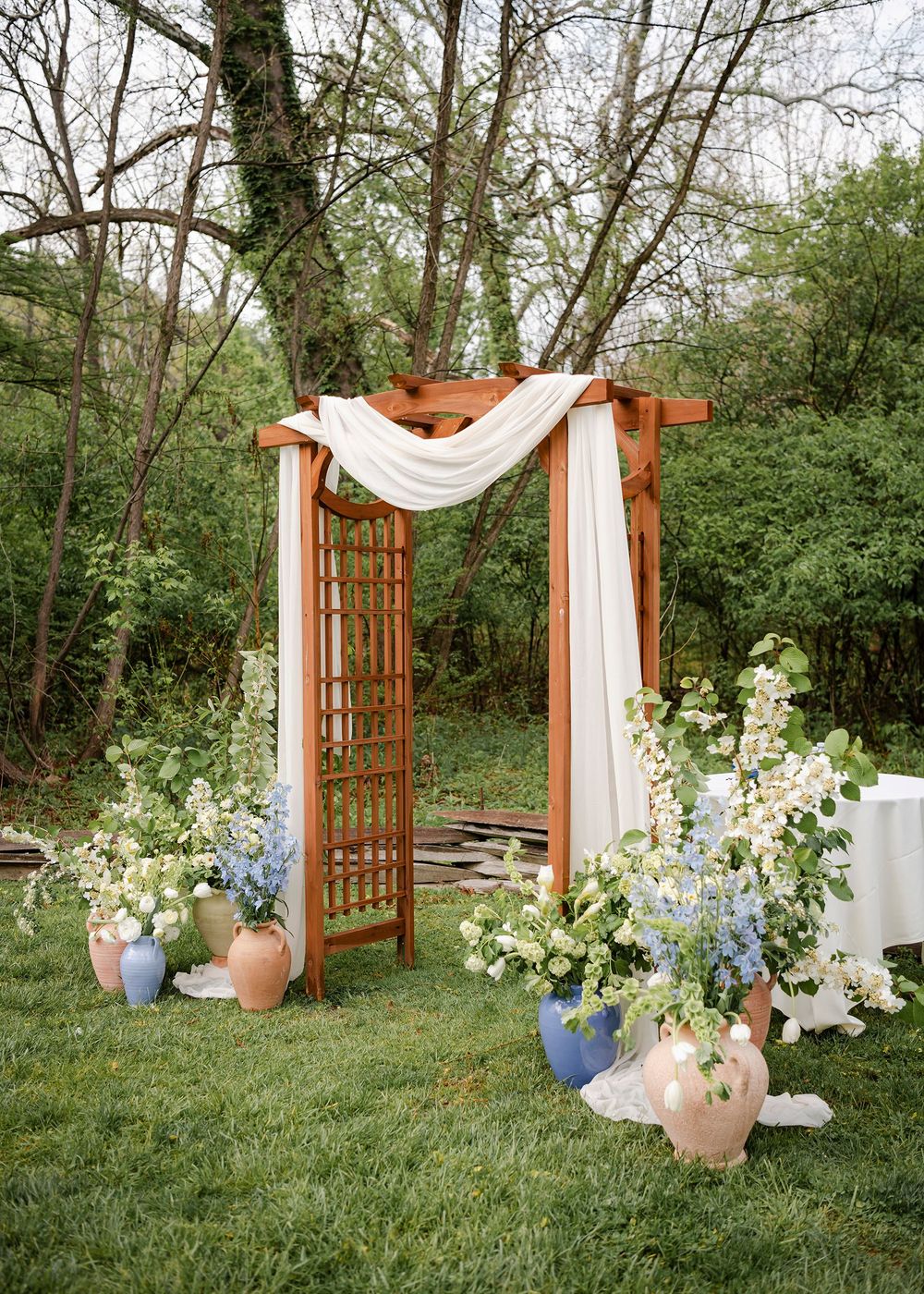 Rustic wooden wedding arch draped with white fabric, adorned with blue and white floral arrangements on green lawn.