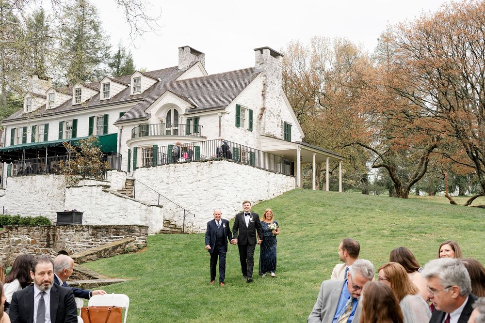Outdoor wedding ceremony on green lawn with white estate house backdrop, guests seated, wedding party walking.