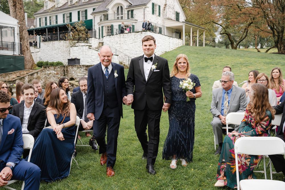 Groom walks down wedding aisle escorted by parents at outdoor ceremony on green lawn near elegant estate.