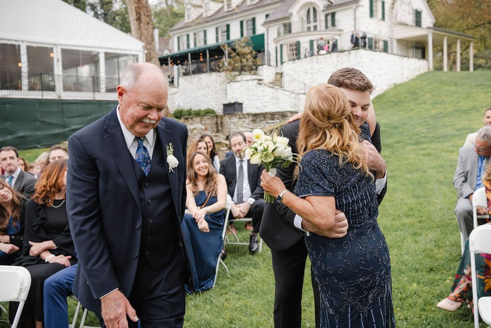 Groom hugging mother at outdoor wedding ceremony while father walks beside them, guests seated on white chairs.