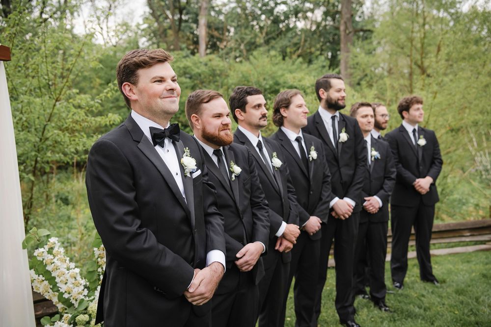 Groom and groomsmen in black tuxedos with white boutonnieres stand outdoors during wedding ceremony.