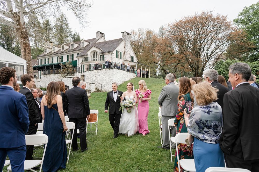 Outdoor wedding ceremony on lush lawn with guests seated in white chairs beside a historic estate venue.