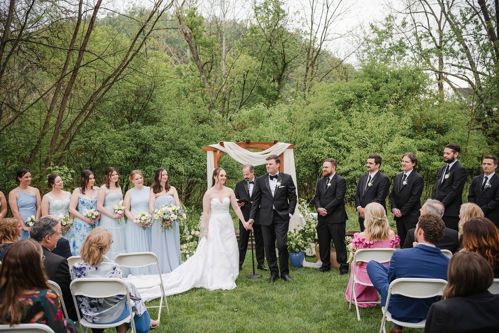 Outdoor wedding ceremony on green lawn with bride, groom, wedding party, and seated guests under a floral arch.