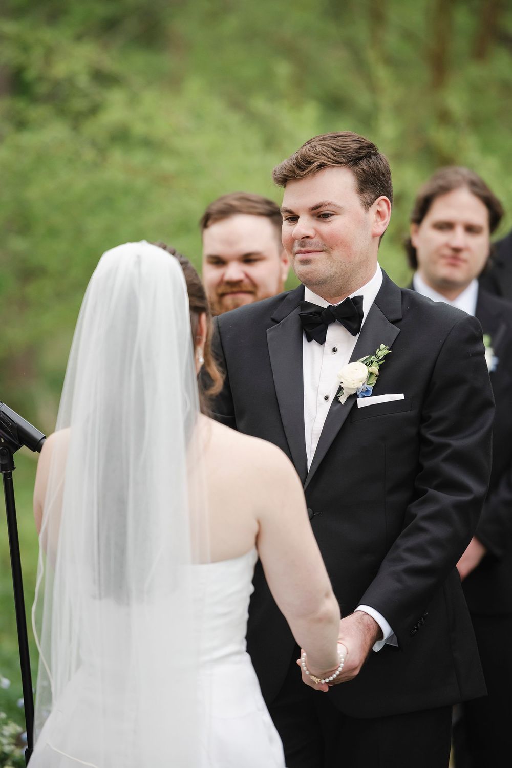 Groom in black tuxedo holds bride's hands at outdoor wedding ceremony with groomsmen standing behind.