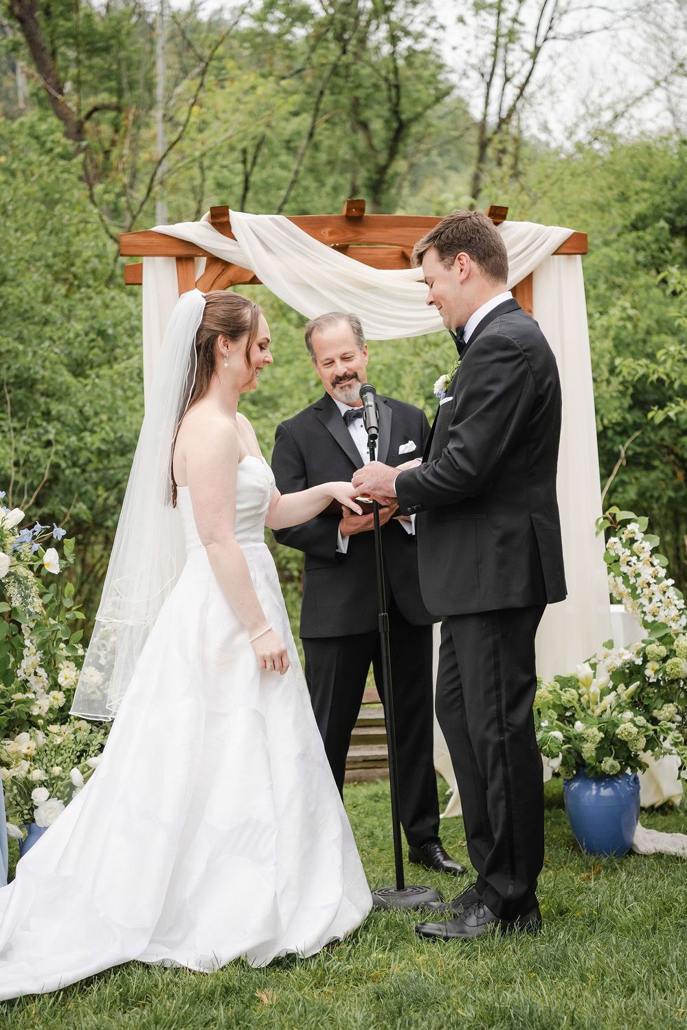 Bride and groom exchange rings outdoors under a draped wooden arch with floral arrangements during a wedding ceremony.