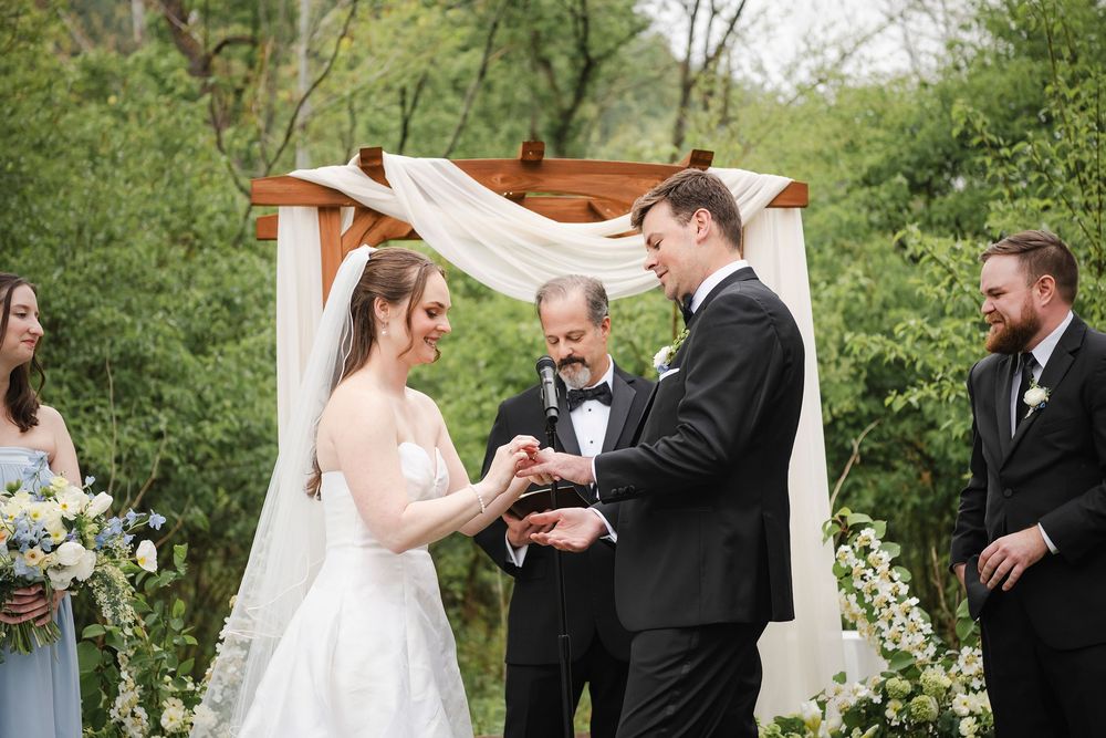 Groom places wedding ring on bride's finger at outdoor ceremony beneath floral arch with officiant and wedding party.