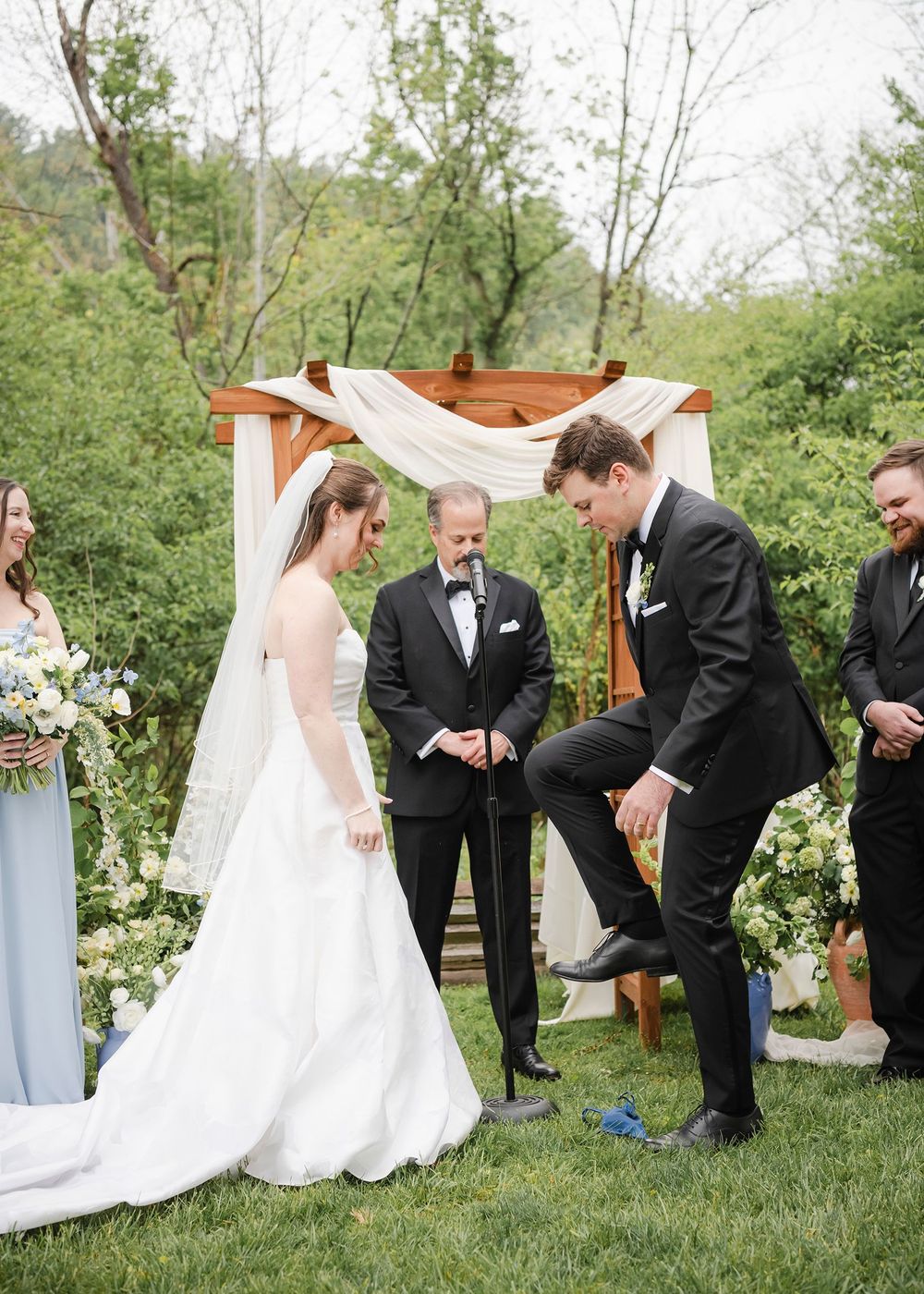 Groom stomps the glass during outdoor Jewish wedding ceremony beneath wooden arch with white draping.