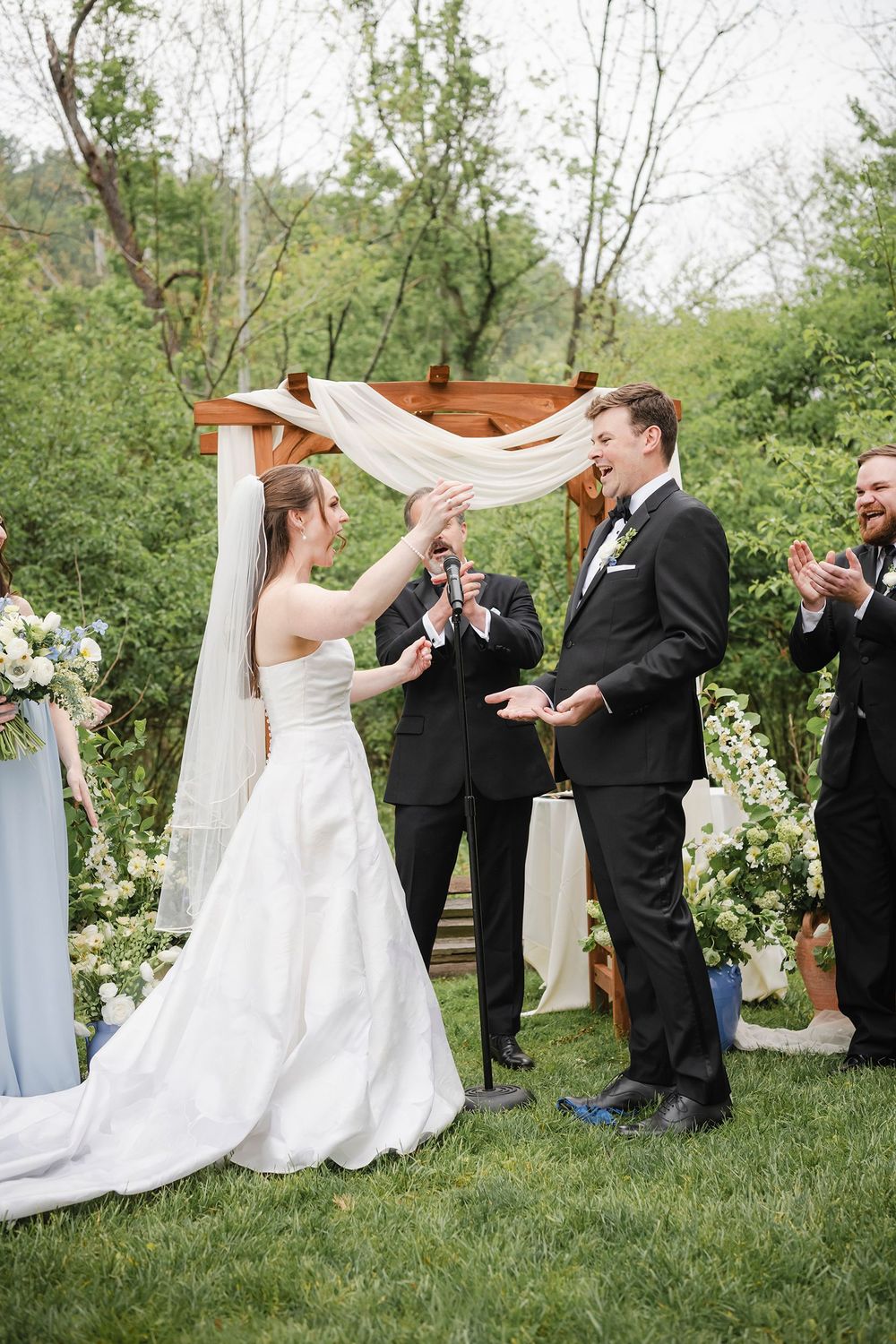 Bride and groom celebrate at outdoor wedding ceremony under wooden arch with white draping and floral arrangements.