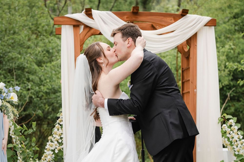 Bride and groom share first kiss under wooden floral arch with white draping at outdoor wedding ceremony.