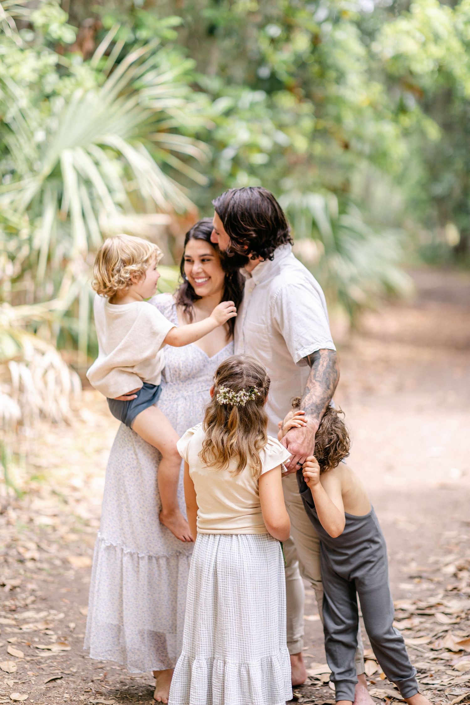 Family of four with tattooed dad kissing mom while children gather around on a tropical garden path.