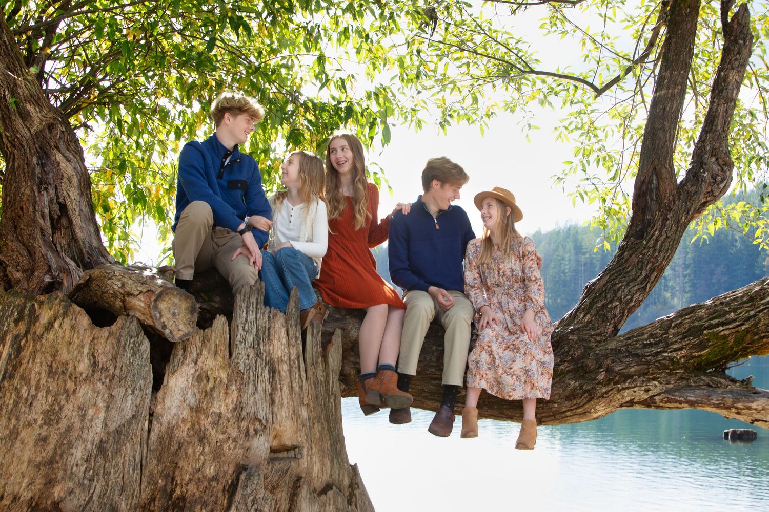 Five children sitting on a large tree branch by a lake, surrounded by lush green trees in natural light.
