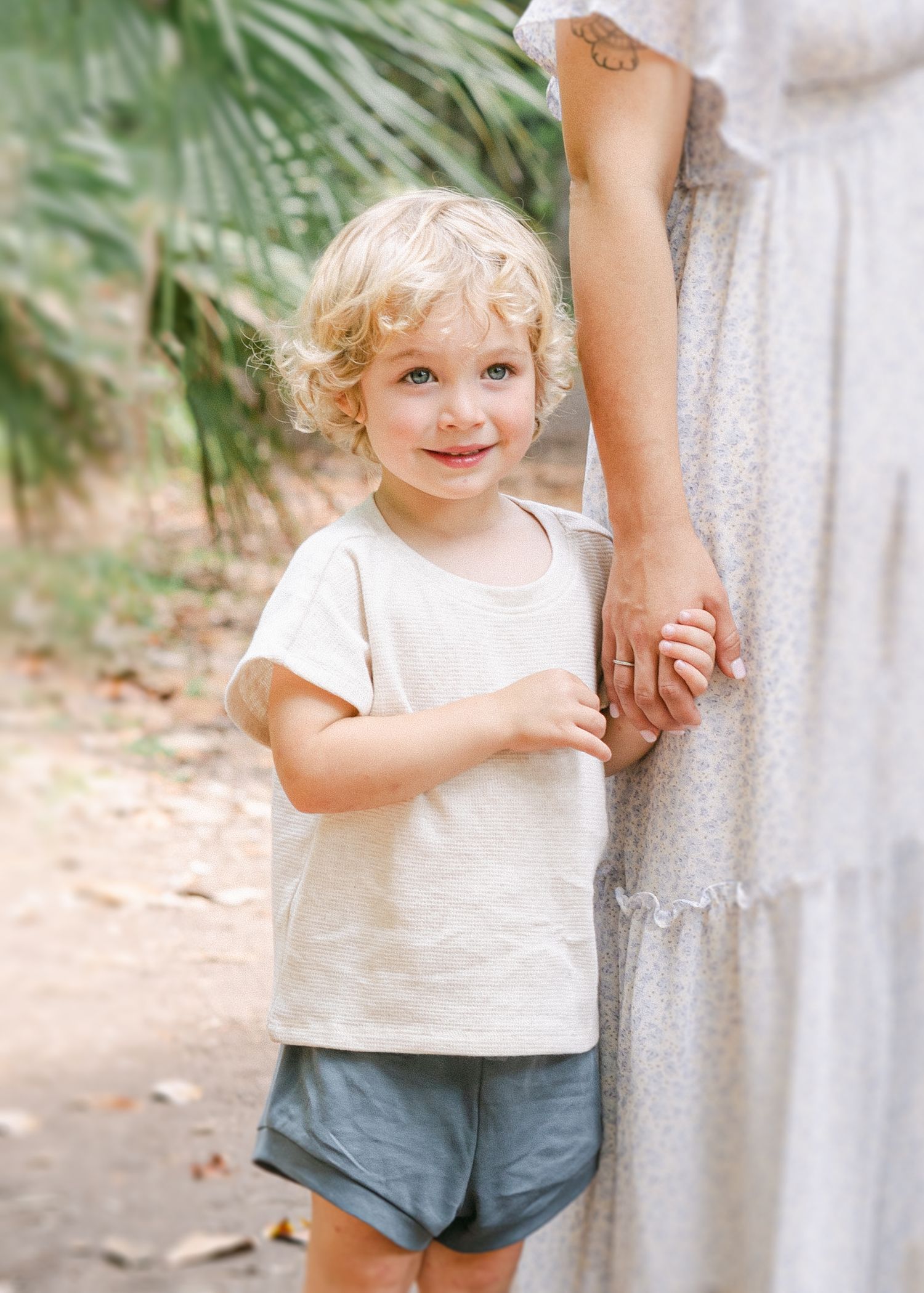 A curly-haired toddler in a cream tee and blue shorts smiles while holding an adult's hand outdoors.