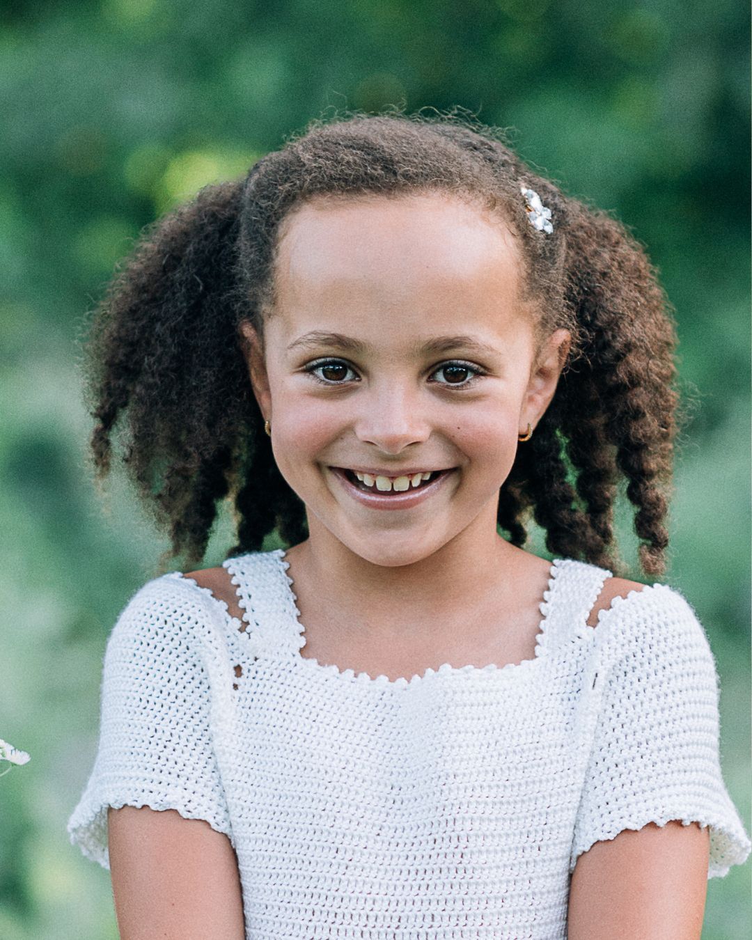 Smiling young girl with curly hair wearing a white crochet top, posing outdoors with green foliage background.
