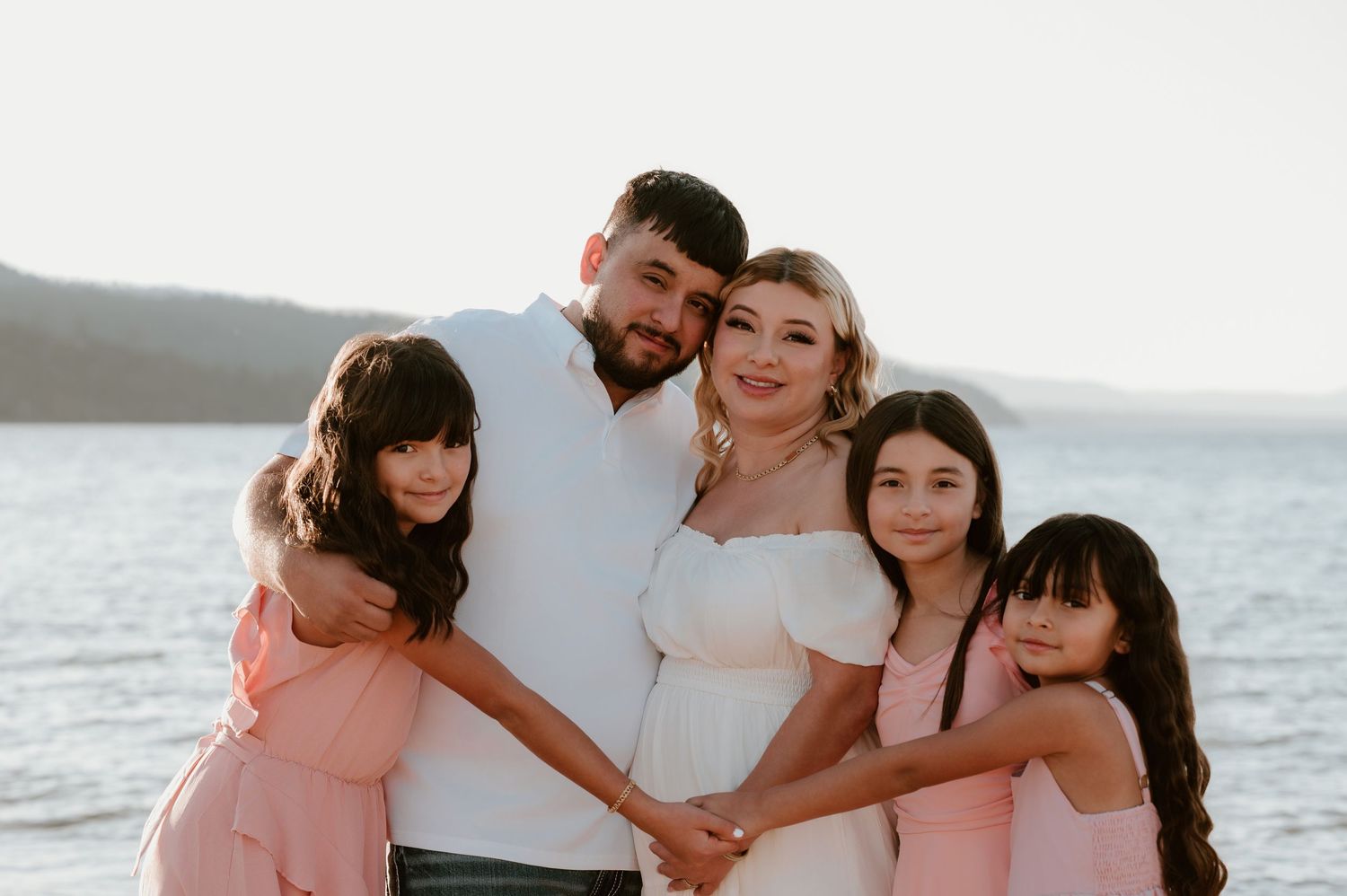 Family of five posing together on a beach, parents with three girls in pink dresses, smiling by the water.