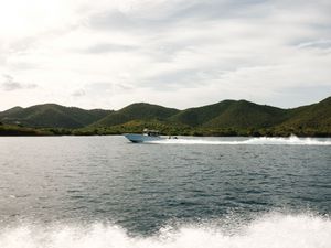 A blue speedboat racing across calm waters with lush green hills in the background under cloudy skies.