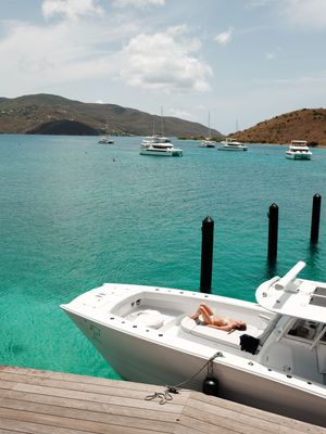 Person sunbathing on a white boat docked at a pier with turquoise waters and sailboats near tropical islands.