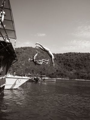 Black and white photo of a person diving off a sailboat into calm water near a forested hill.