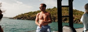 Shirtless man in blue swim trunks and sunglasses relaxing on a boat with tropical green hills and calm water behind him.
