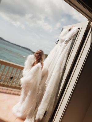 Bride in white fur wrap views her wedding gown hanging by a balcony with ocean views.