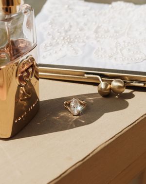 Gold ring with crystal stone on a surface beside a rose gold perfume bottle and pearl-embellished clutch.