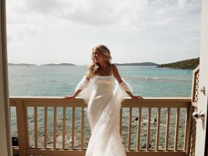 Bride in white gown and fur wrap poses on balcony overlooking turquoise Caribbean waters.