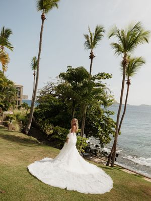 Bride in a stunning white gown with long train stands on tropical lawn overlooking the ocean with palm trees.