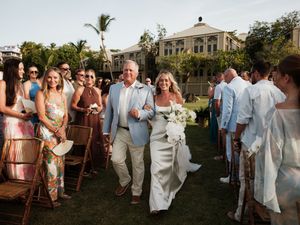 Bride walks down outdoor wedding aisle with escort amid smiling guests on tropical lawn.