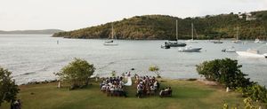 Outdoor waterfront wedding ceremony on green lawn with sailboats anchored in tropical bay surrounded by lush hills.