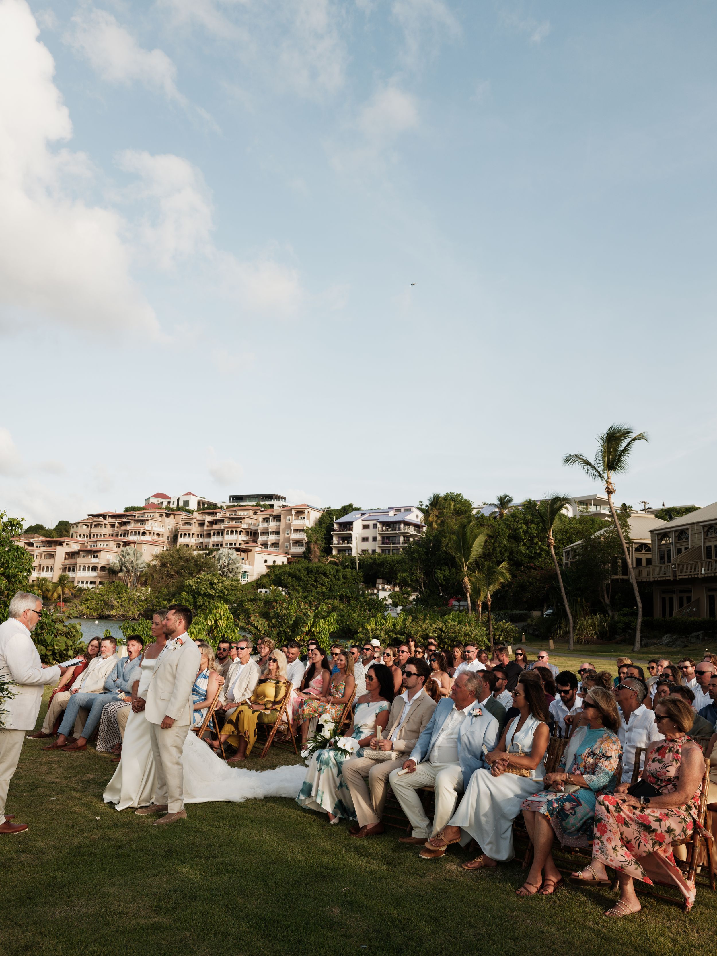 Outdoor tropical wedding ceremony with seated guests on lush lawn overlooking hillside resort.