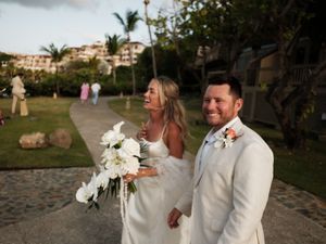 Happy newlywed couple walks together outdoors, bride holding white bouquet, groom in white suit at tropical venue.
