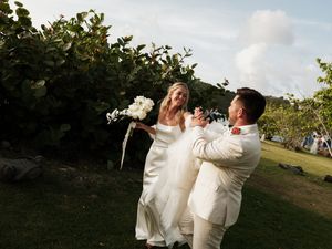 Joyful bride and groom share a playful moment on tropical lawn, bride holding white bouquet in white wedding attire.