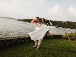 Groom lifts bride in white dress on lush lawn by turquoise bay at tropical waterfront wedding.