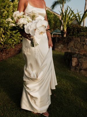 Bride in elegant satin gown holds white orchid bouquet in lush garden setting.