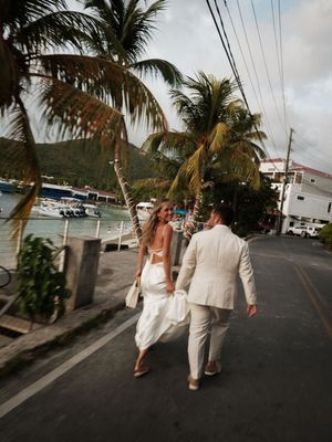 Bride and groom walking hand in hand along a tropical waterfront street lined with palm trees.