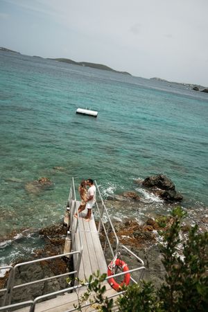Couple kissing on a wooden dock above rocky turquoise Caribbean waters with a boat in the distance.