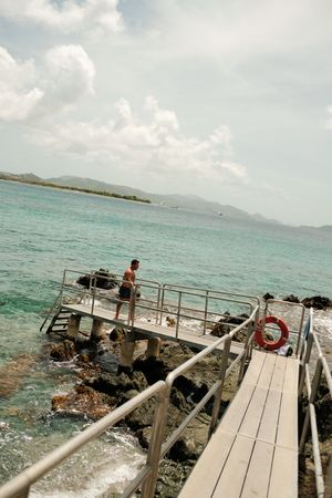 Man standing on a rocky ocean pier with turquoise Caribbean waters and mountainous islands in the background.