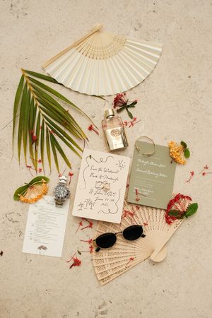 Tropical wedding flat lay with fans, invitations, perfume, sunglasses, and floral details on stone surface.