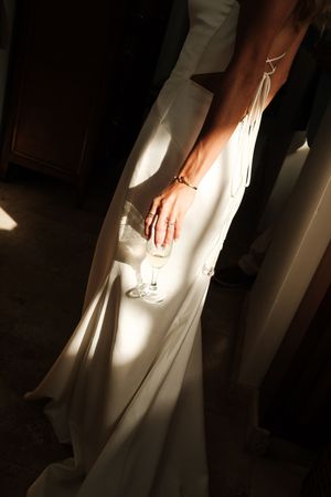 Bride in white satin gown holding champagne glass with sunlight casting dramatic shadows.