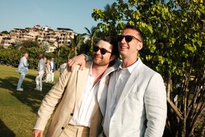 Two men in light suits smiling and embracing outdoors at a resort event on a sunny day.