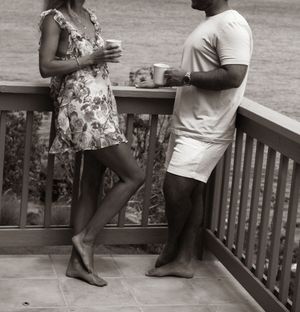 Couple in casual attire enjoying drinks together on a wooden balcony overlooking greenery.