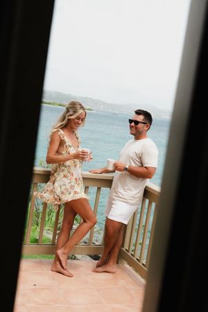Couple enjoys morning coffee on oceanview balcony, woman in floral dress and man in casual white outfit.