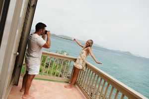 Man photographs woman posing on tropical oceanview balcony, both in casual summer attire with scenic water views.