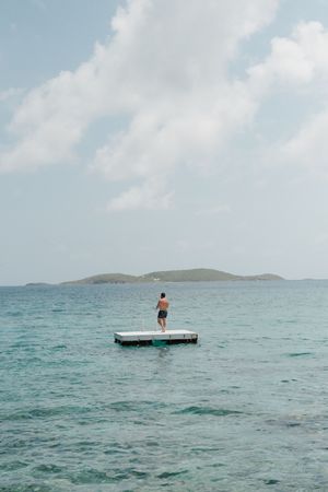 Man stands alone on floating dock platform surrounded by turquoise Caribbean waters with island in the background.