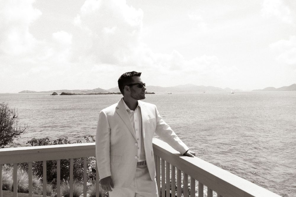 Elegant man in white suit stands on seaside balcony, gazing at ocean horizon in black and white photo.