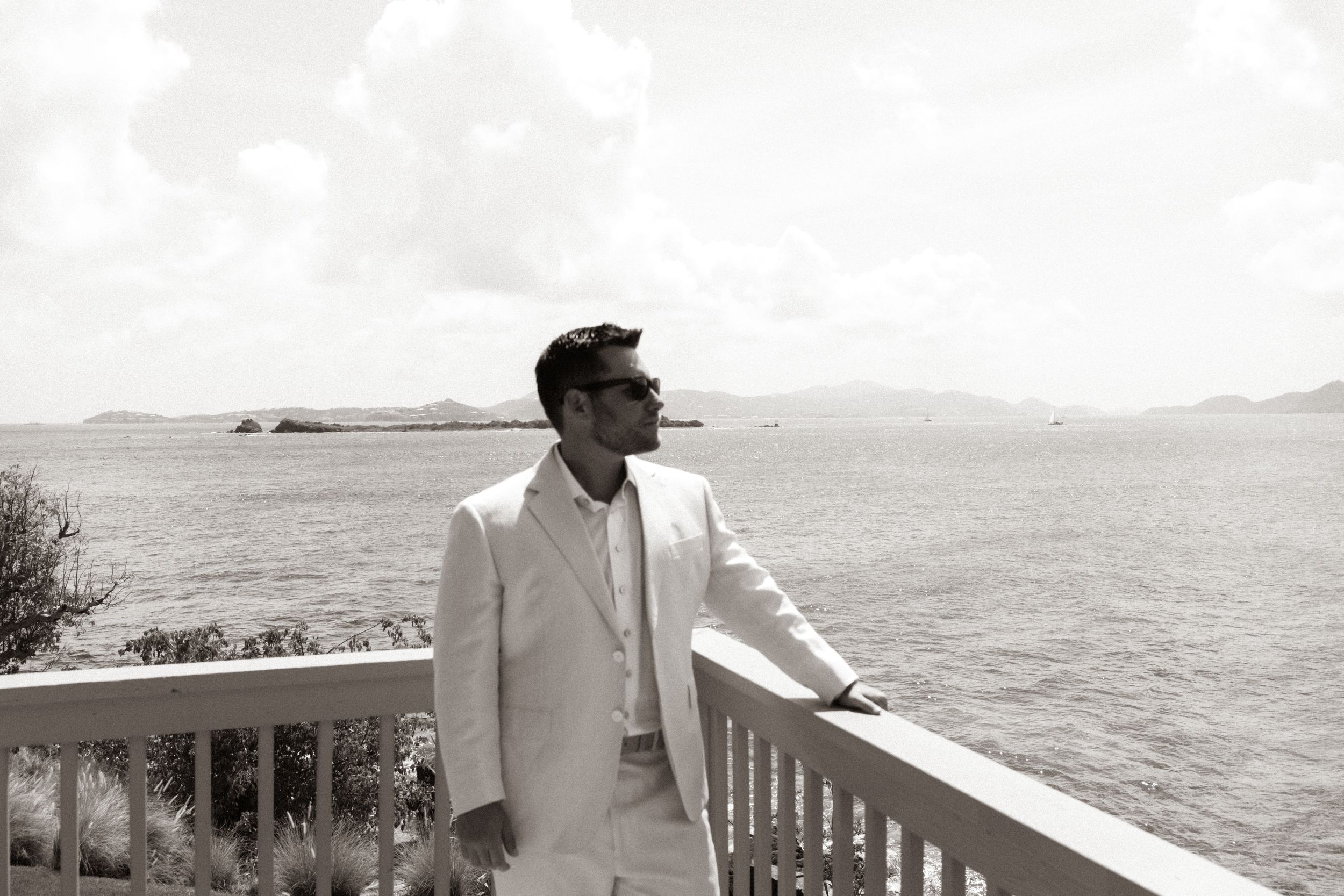 Elegant man in white suit stands on seaside balcony, gazing at ocean horizon in black and white photo.
