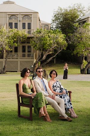 Three elegantly dressed people sit on a wooden bench on a green lawn with a luxury building backdrop.