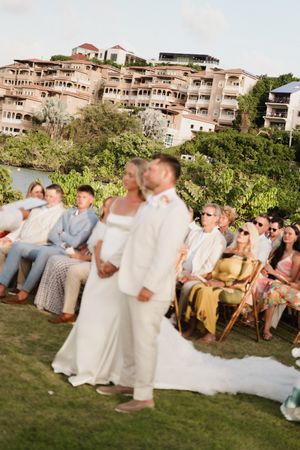 Bride and groom stand at outdoor ceremony with resort backdrop and seated guests.